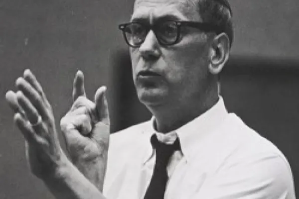 Black and white depiction of a man in glasses caught midspeech, gesturing with his hands