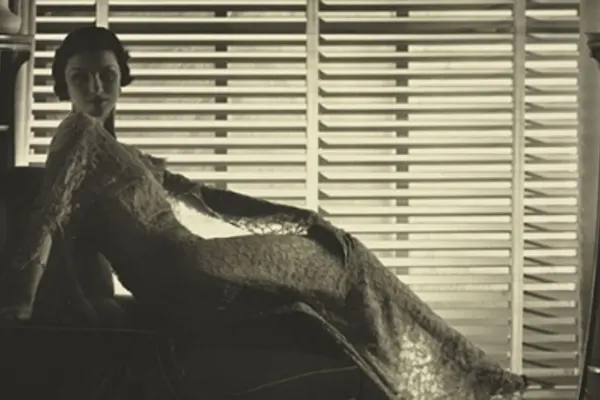 A woman in an elegant, lacy gown lounges against an armchair in front of a wall of windows covered in blinds. Her body is profile but her face is turned towards the camera. Everything is in sepia tones.