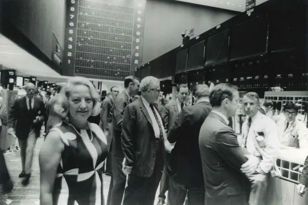 Woman in print dress standing on Stock Exchange floor surrounded by men