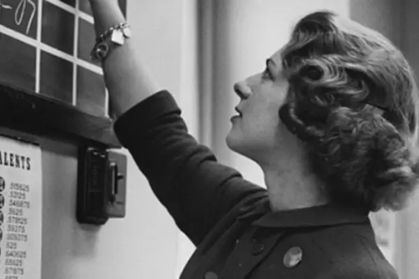 A woman with pin-curled hair stands profile, writing on a blackboard hung on the wall. Everything is in black and white.