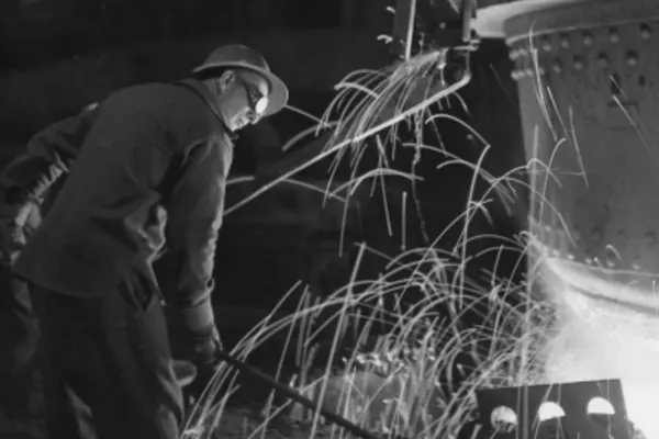 A goggle wearing steel worker at work beside a large steel contraption emitting sparks.