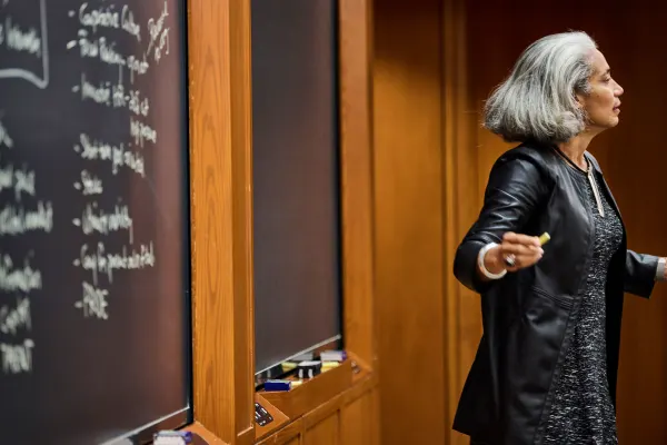 A professor engages an out-of-frame classroom with a blackboard full of writing behind her