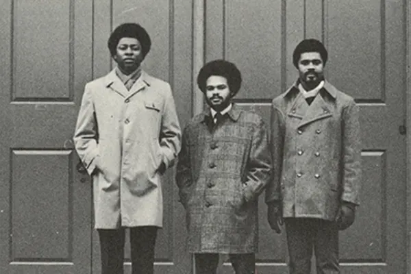 Three young black men in coats stand before a set of doors, facing forward.