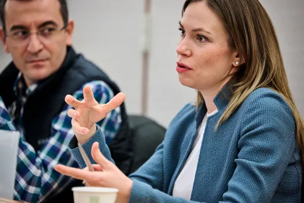 Close up of a woman in a blue blazer speaking in a classroom. A man in a plaid shirt and black vest with glasses listens attentively