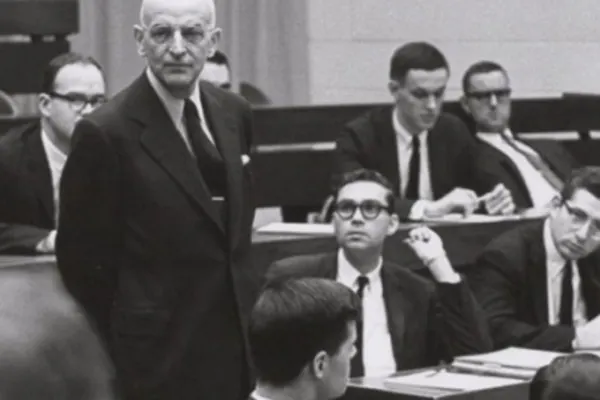 A single man in a suit faces forward, standing before a group of seated men in suits. Those who are seated have pens and paper before them. One looks directly up at the standing man.