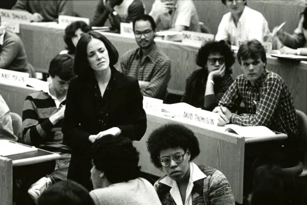 A woman stands and gazes out thoughtfully amidst a lecture hall of sitting students