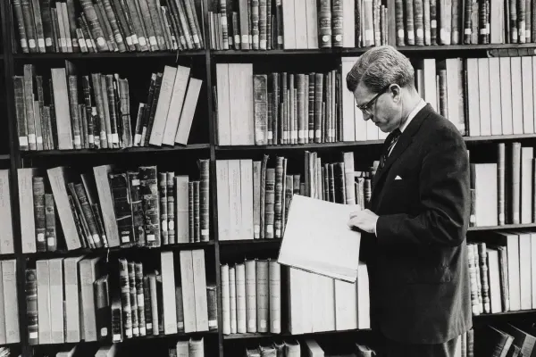 A man stands before large bookshelves. He is reading a book that lies open in his hands.