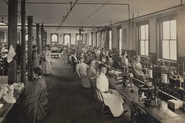 Women sit in rows at worktables with sewing machines. Some are looking at the camera, while others are looking at their work.