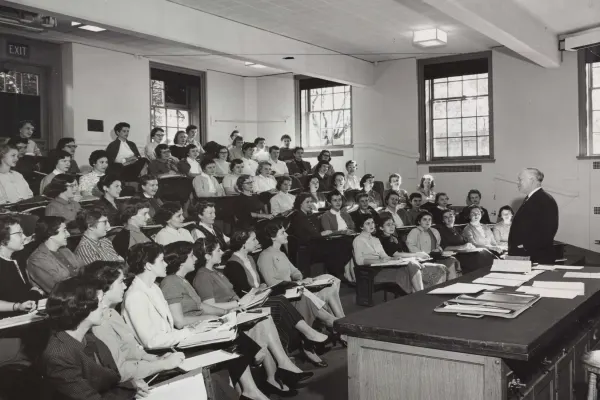 A classroom of young women sit attentively listening to a lecturer who stands before a table strewn with various papers.