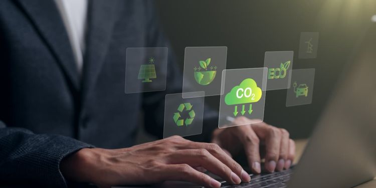 A business professional typing on a laptop with icons floating above it, including a solar panel, a recycling symbol, Earth with plants sprouting from it, CO2, the word "Eco," an electric vehicle, and solar panels