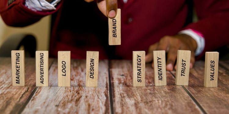 A business professional placing wooden blocks with text on a table. The one they're holding reads "brand," and the others read "marketing," "advertising," "logo," "design," "strategy," "identity," "trust," and "values."