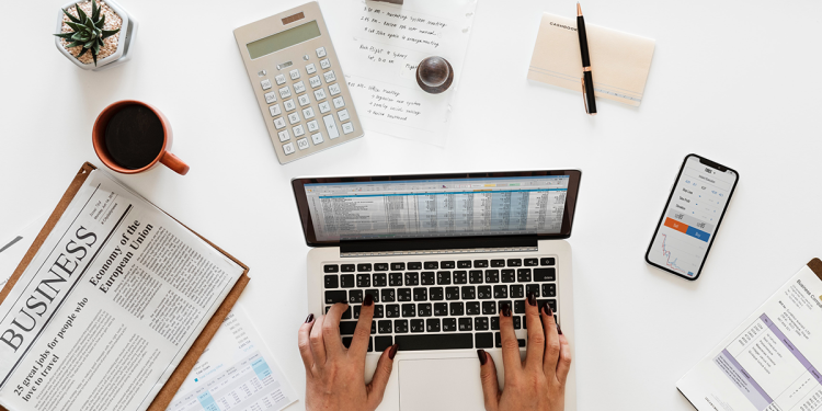 Woman typing on a laptop surrounded by papers, calculator, and phone