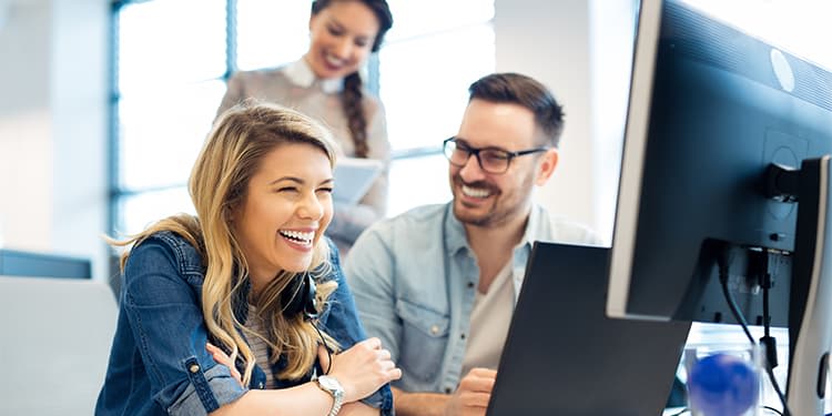 Two employees working collaboratively on a computer, with another employee smiling, taking notes behind them.