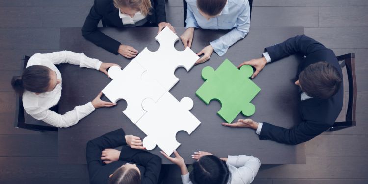 Business team seated at table fitting large puzzle pieces together (three white and one green)