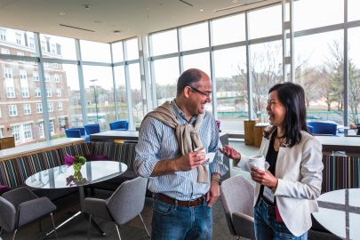 A male and female executive smiling and talking whle drinking coffee and standing in a lounge area.