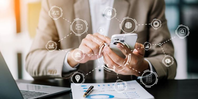 Business professional using a smartphone with icons floating around it, including a graph, mail envelope, and lock. A pile of business charts are on the desk in front of them.