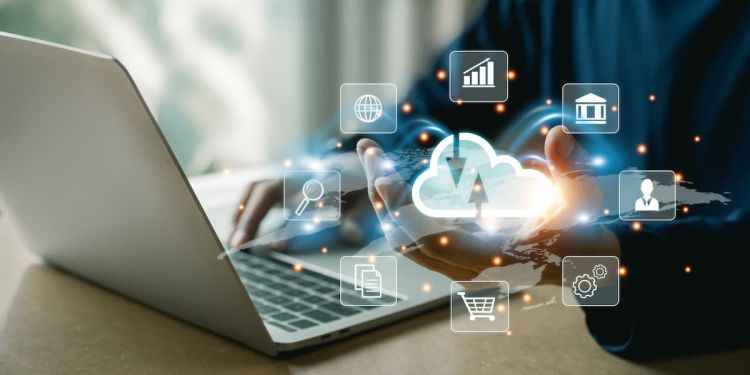 Person seated at laptop with icons floating around their hand, including a bar graph, building, business professional, gears, shopping cart, documents, magnifying glass, and globe