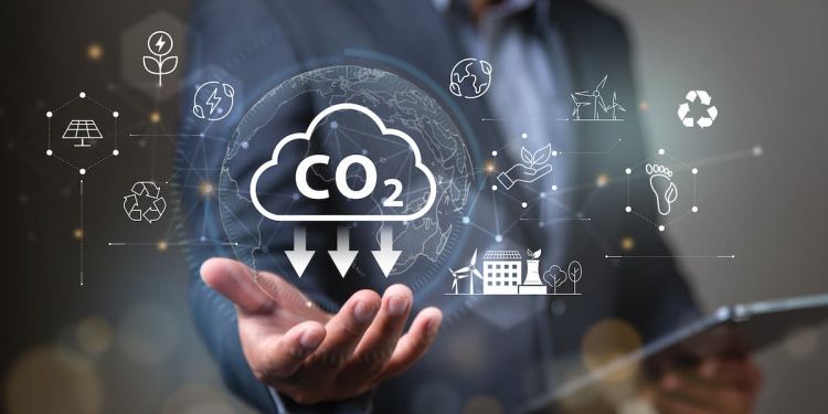Business professional holding a tablet with an icon signifying reducing carbon emissions above their outstretched hand. Other icons are scattered around the image, including a solar panel, windmills, and Earth.