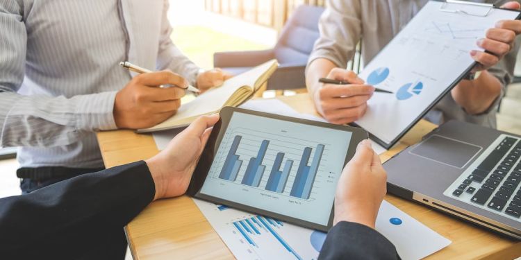 Three people sitting at a table. Two of them are comparing different graphs and the third person is taking notes.